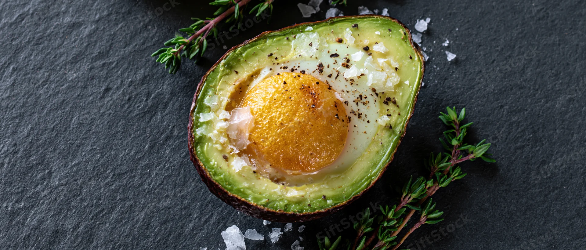 Baked egg in halved avocado on slate with thyme and flaky salt