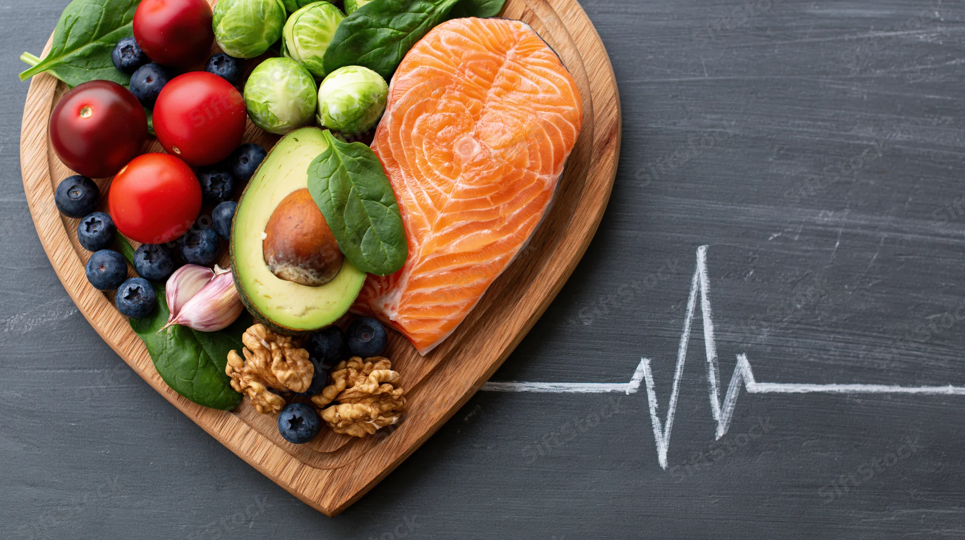 Heart-shaped wooden board with salmon, avocado, berries, nuts and greens beside a chalk-drawn heartbeat on dark surface