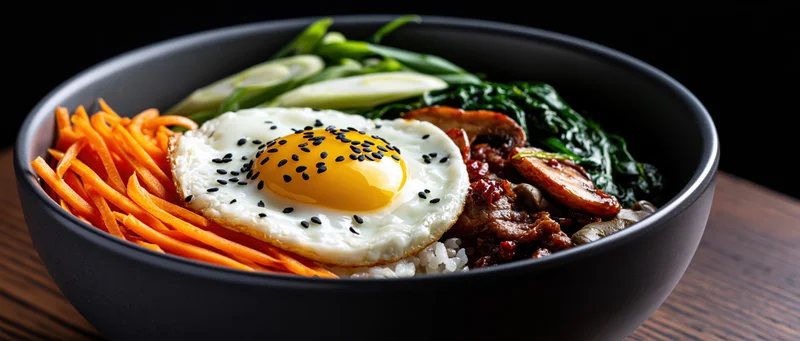 Korean bibimbap in a dark matte ceramic bowl with fried egg, carrots, spinach, mushrooms and meat on wooden surface