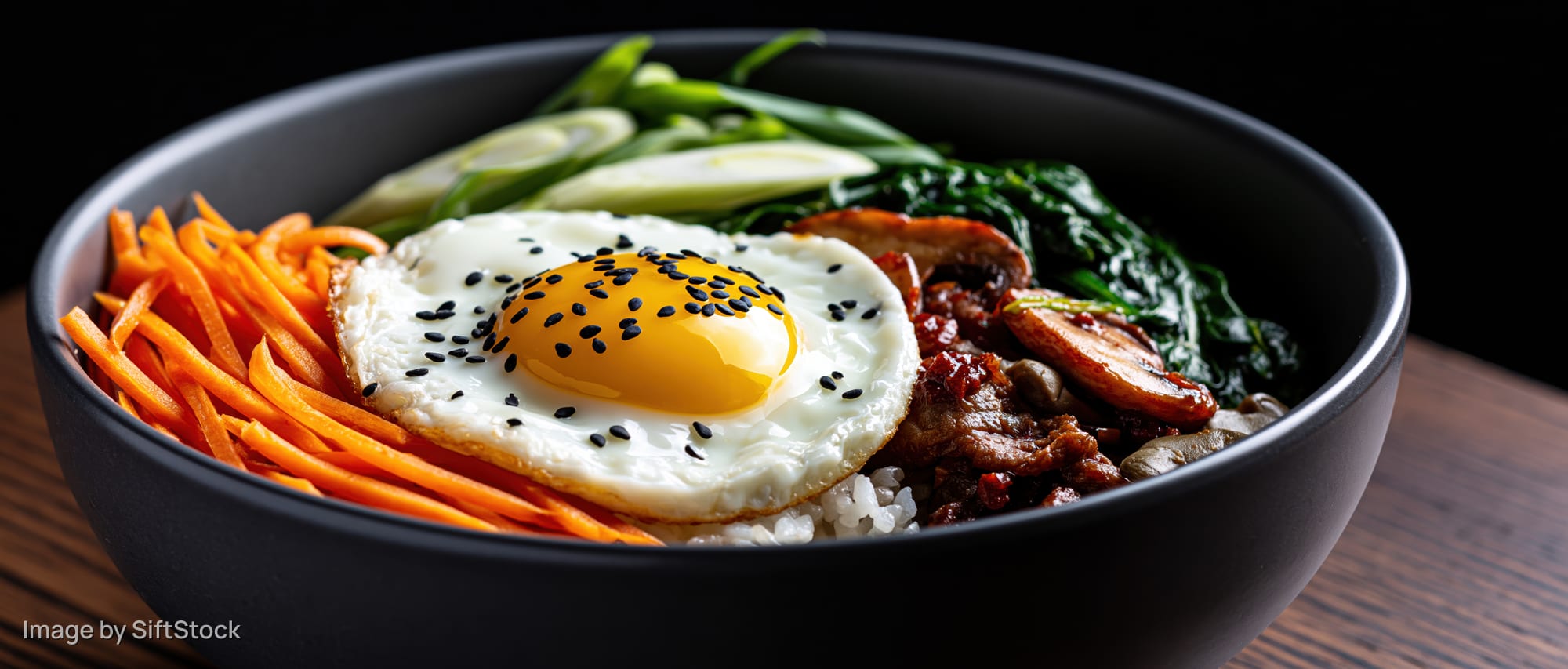 Korean bibimbap in a dark matte ceramic bowl with fried egg, carrots, spinach, mushrooms and meat on wooden surface