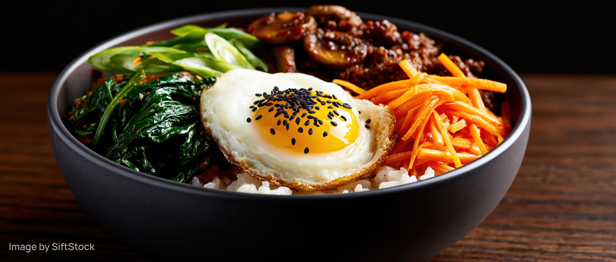 Korean bibimbap in a dark matte ceramic bowl with rice, vegetables, beef, and a fried egg topped with sesame seeds on a wooden table