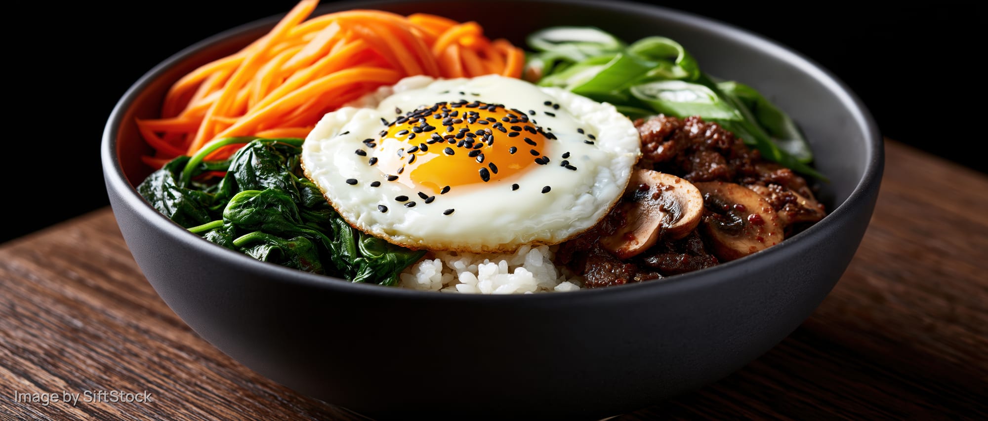 Korean bibimbap in a dark matte ceramic bowl with fried egg and vegetables on wooden surface