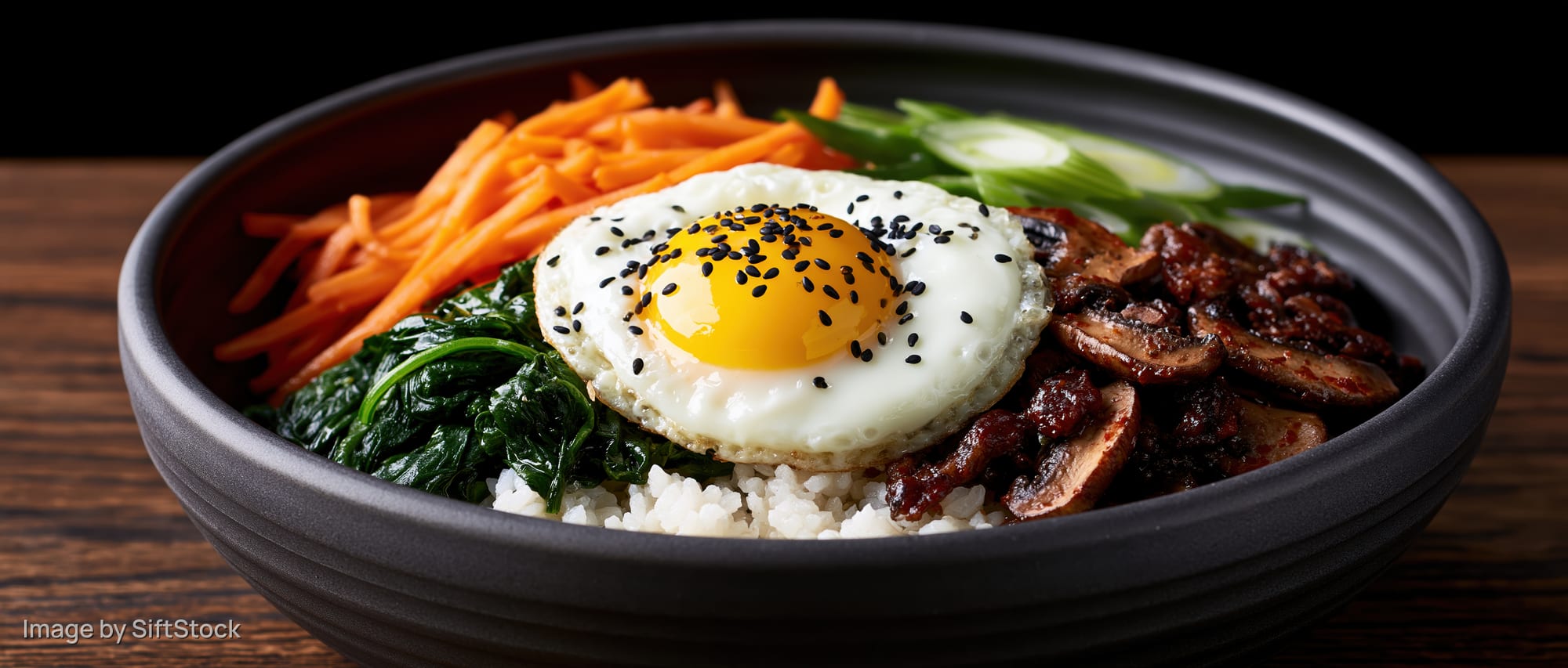Korean bibimbap in a dark matte ceramic bowl with rice, vegetables, beef, and a fried egg topped with sesame seeds on a wooden table