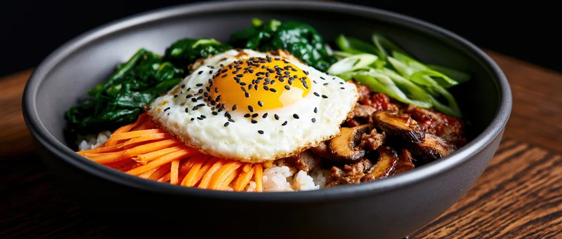 Korean bibimbap in a dark matte ceramic bowl with fried egg, vegetables and beef on wooden surface