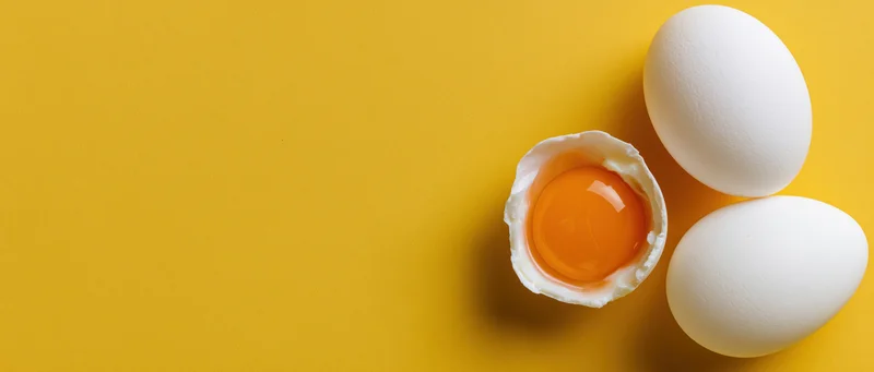 Cracked egg half with yolk and two whole eggs on yellow surface