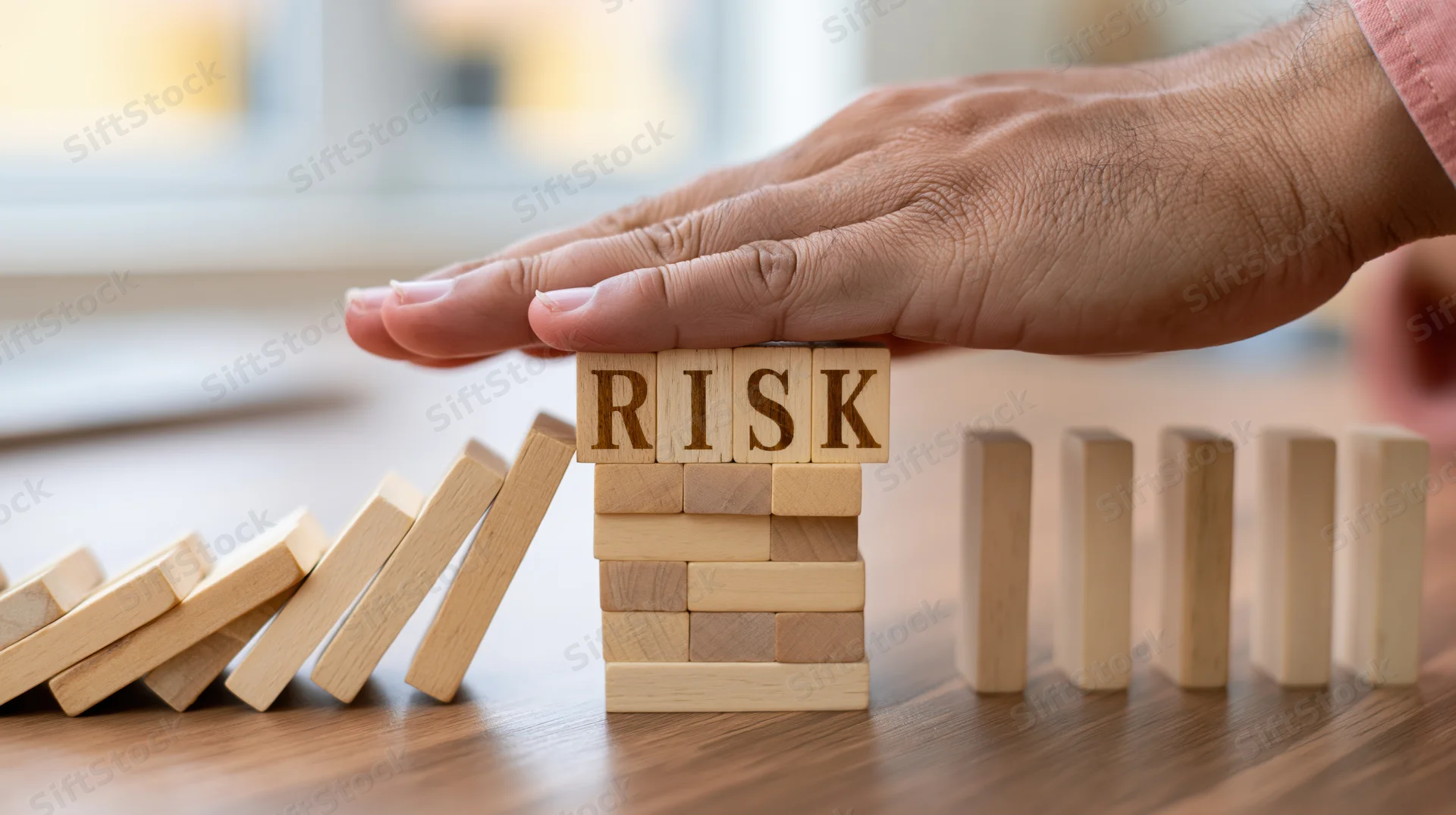 Hand stopping wooden blocks spelling risk in a domino chain on a desk