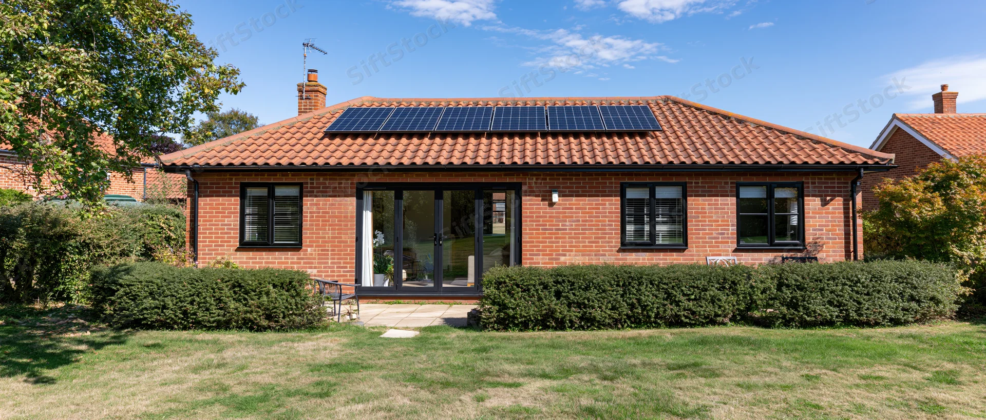 Red brick house with solar panels on terracotta roof surrounded by garden and hedge