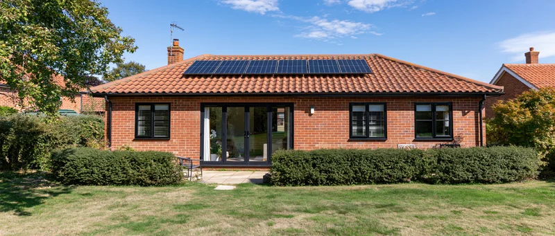 Red brick house with solar panels on terracotta roof surrounded by garden and hedge