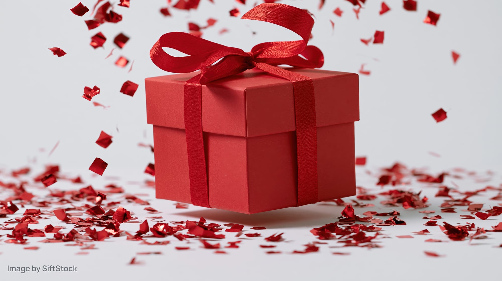 Red gift box with ribbon and bow surrounded by falling confetti on white background