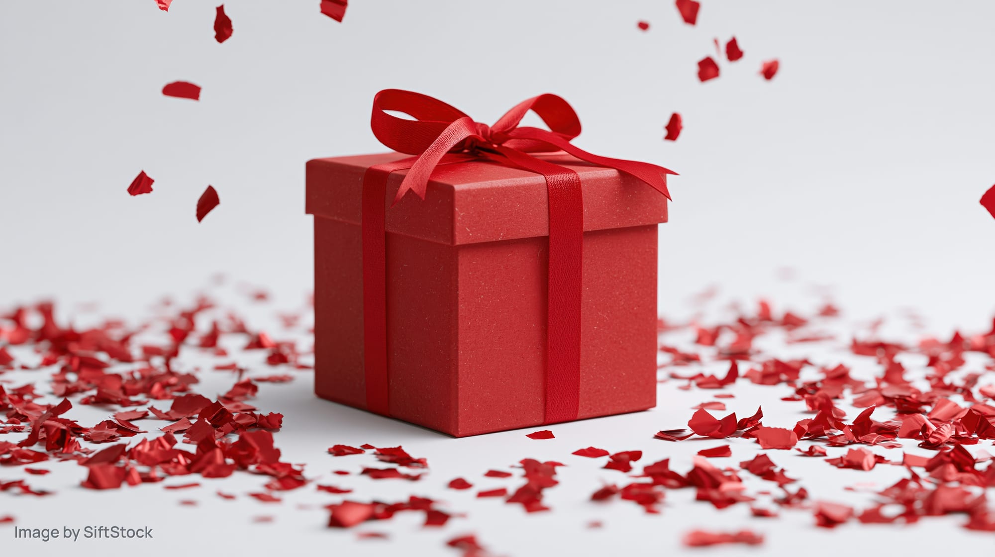 Red gift box with ribbon and bow surrounded by falling confetti on white background