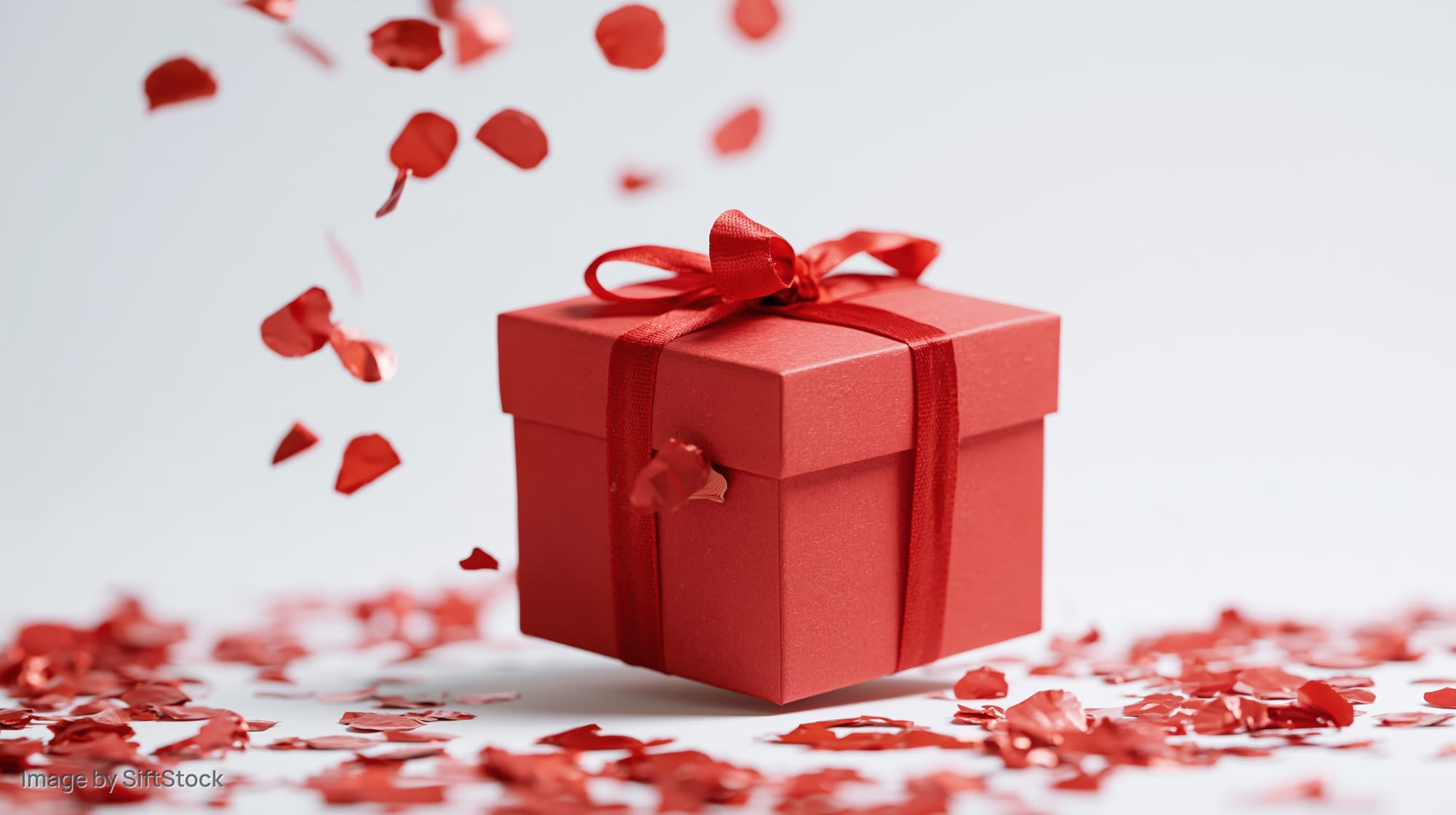 Red gift box with ribbon and bow surrounded by falling red confetti on white surface