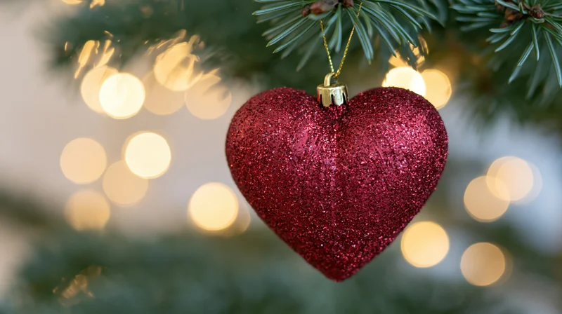 Red glitter heart ornament hanging from a pine tree branch with blurred holiday lights in the background