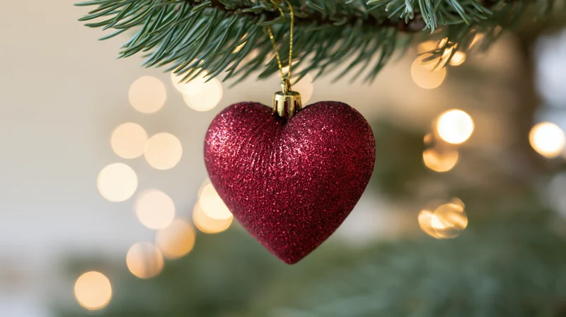 Red glitter heart ornament hanging from a pine tree branch with blurred holiday lights in the background