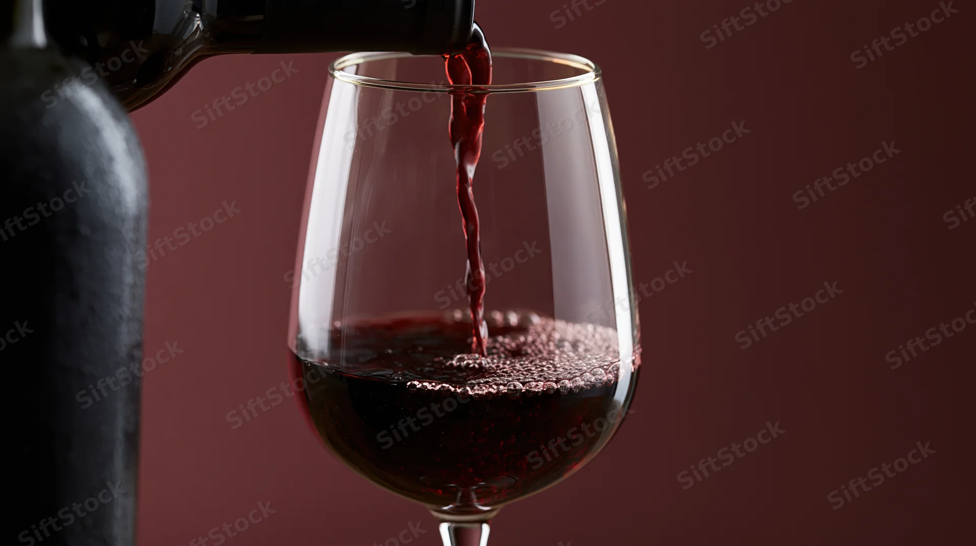 Red wine being poured from a dark bottle into a clear stemless glass against a deep maroon background