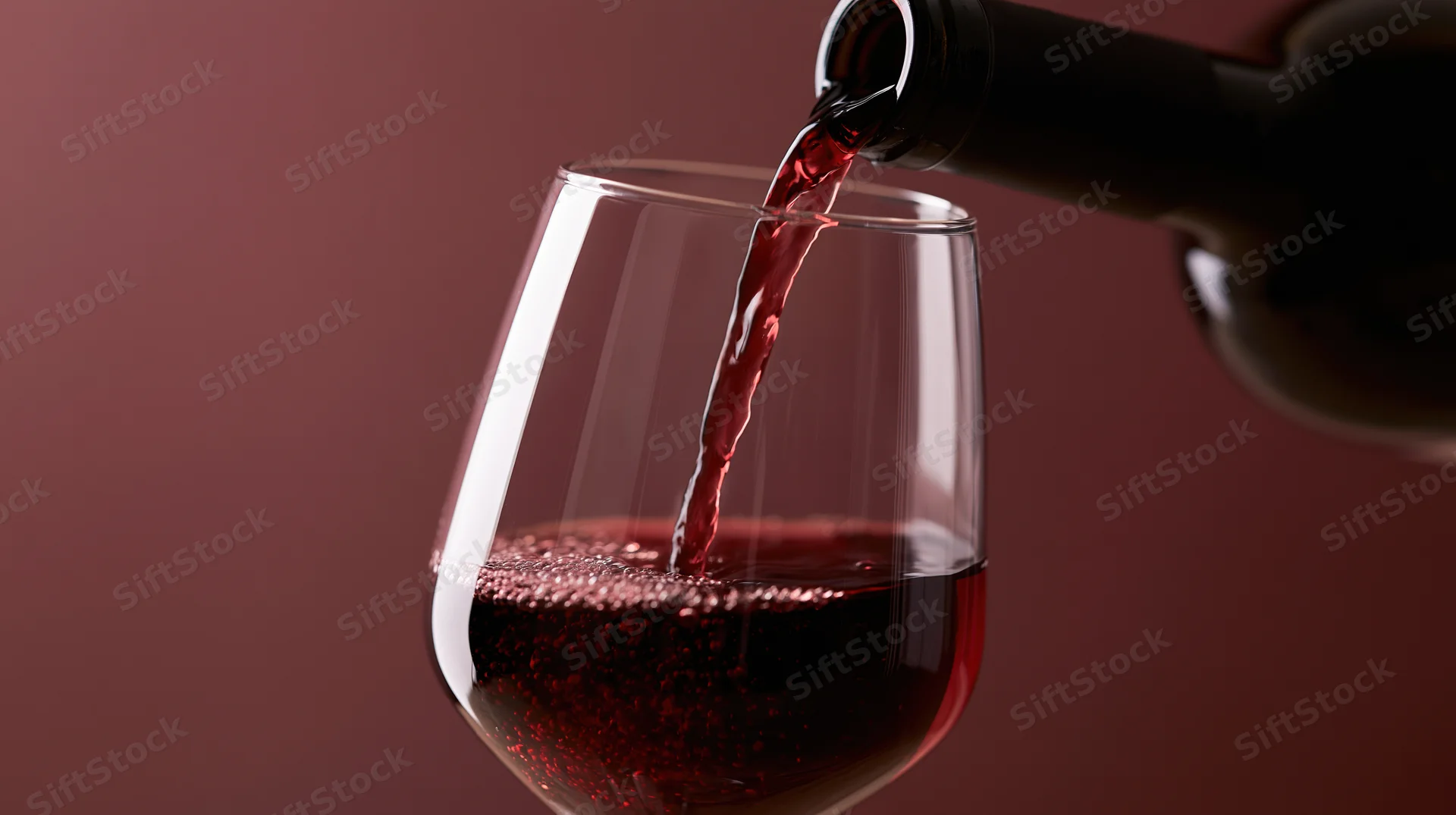 Red wine being poured from a dark bottle into a clear stemless glass against a maroon background