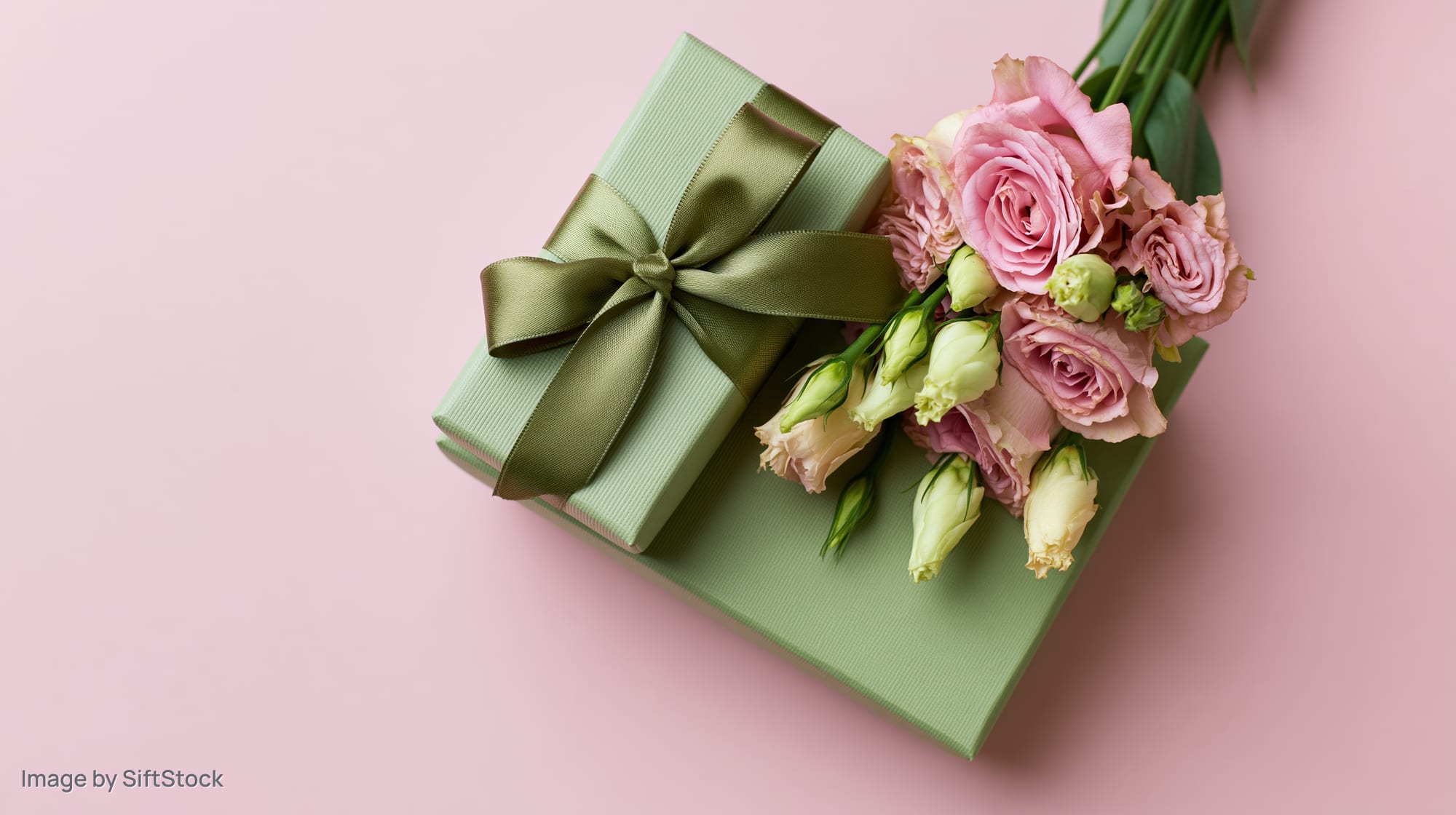 Sage green gift box with satin ribbon bow beside a bouquet of pink roses on a pastel pink surface