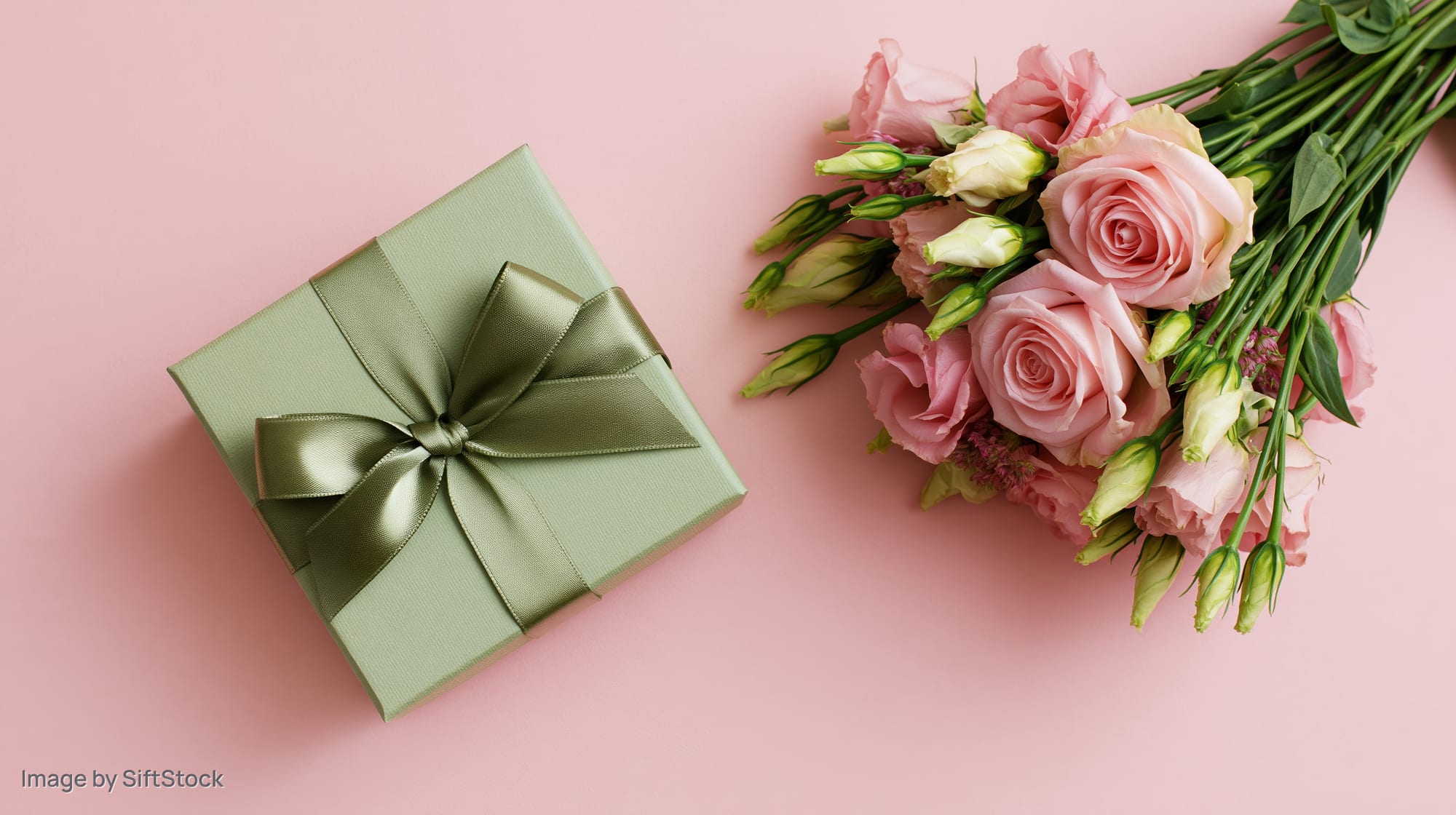 Sage green gift box with satin ribbon bow beside a bouquet of pink roses on a pastel pink surface