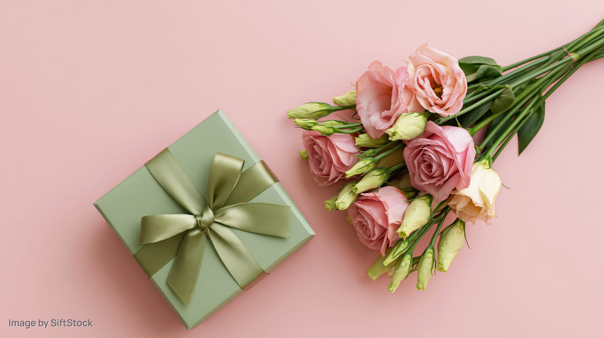 A sage green gift box with a satin ribbon bow beside a small bouquet of pink roses on a pink surface