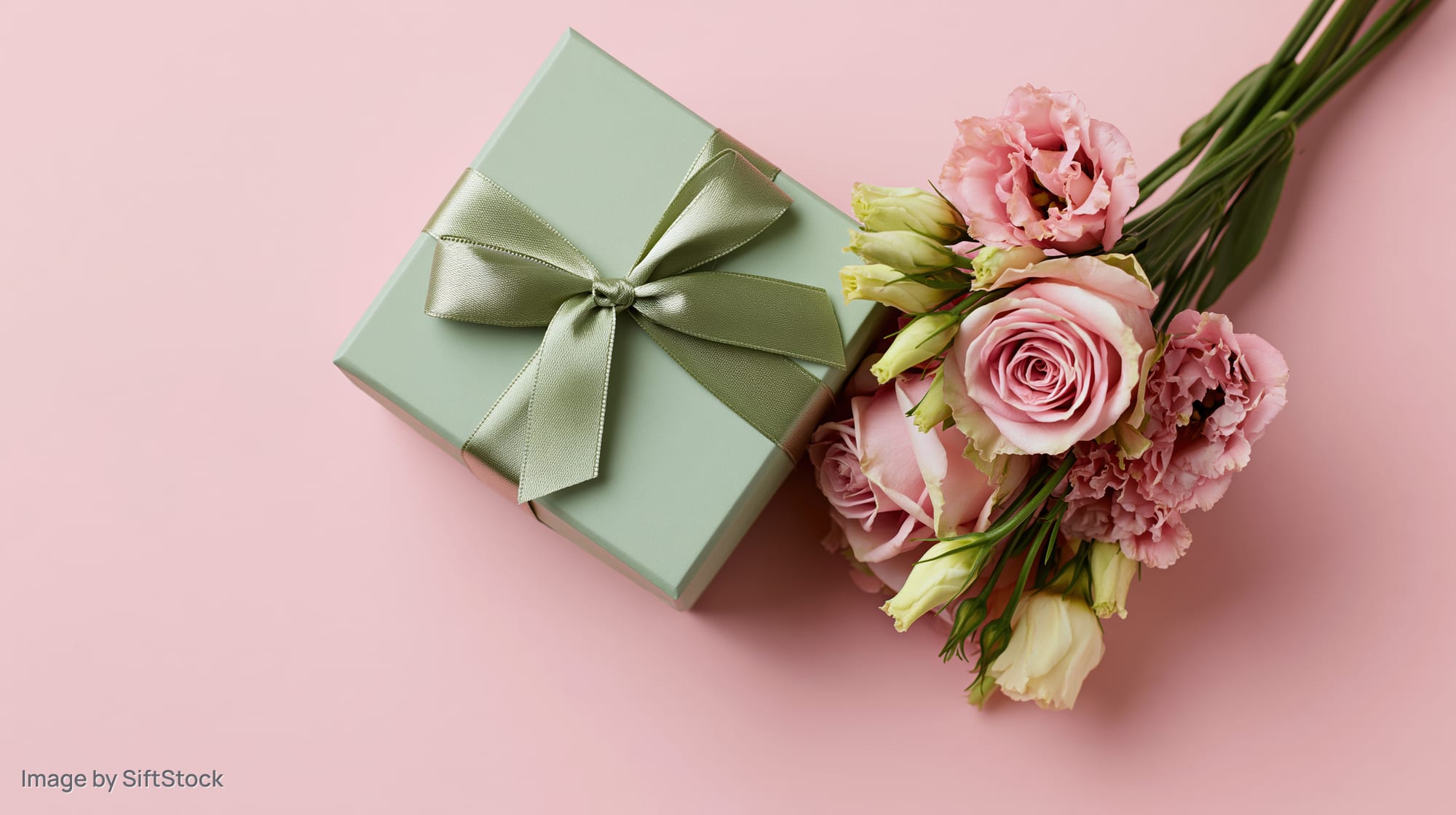 Sage green gift box with satin ribbon bow beside a bouquet of pink roses and carnations on a pink background