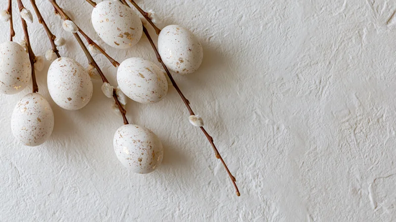 White speckled easter eggs with gold foil accents arranged on a textured white surface with willow branches and catkins, flat lay composition
