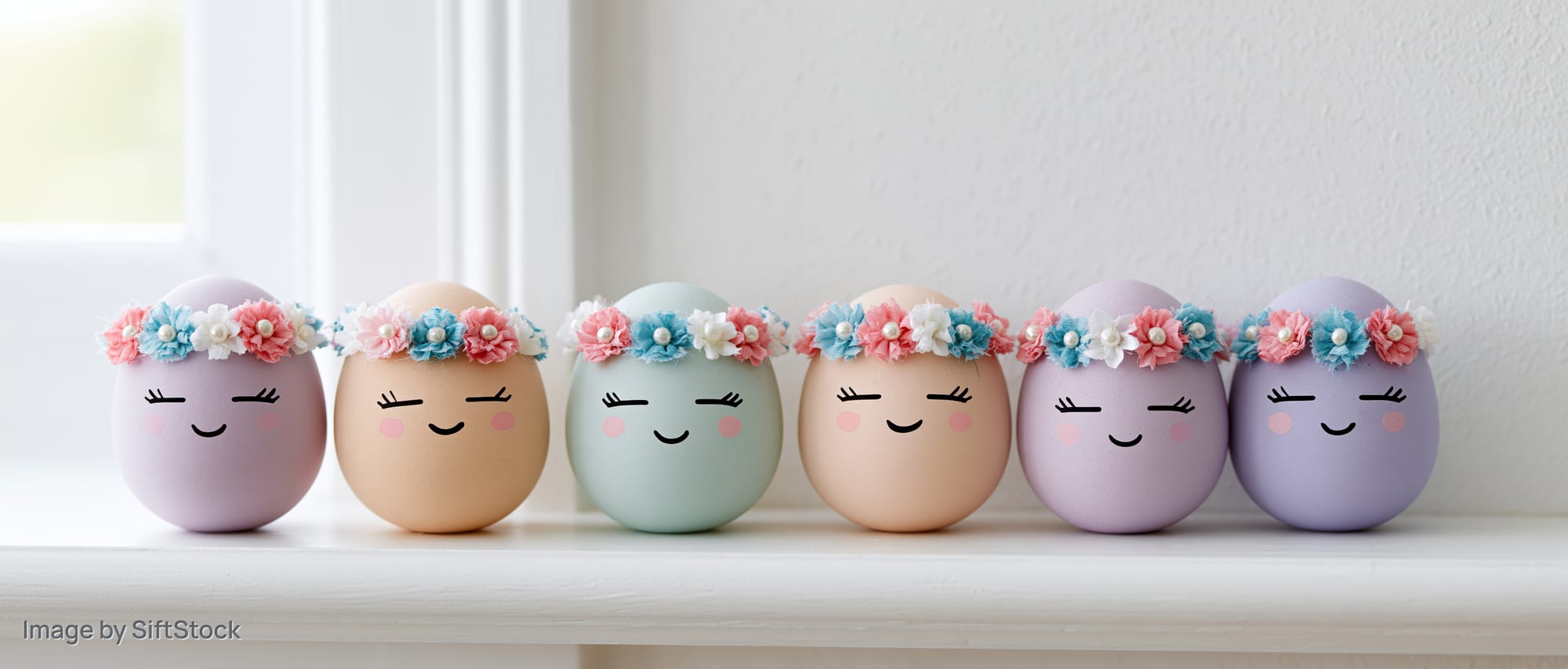 Six pastel easter eggs with cartoon faces and floral crowns arranged in a row on a windowsill