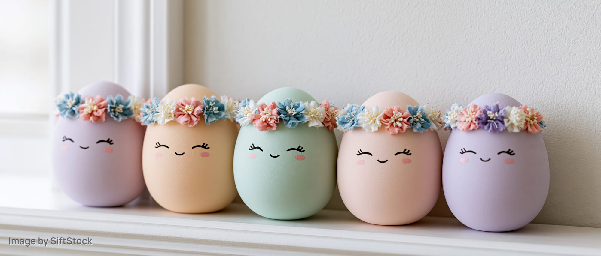 Six pastel easter eggs with cartoon faces and floral crowns arranged in a row on a windowsill