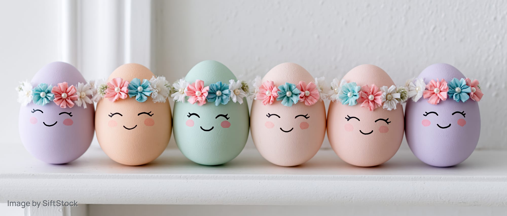 Six pastel-colored Easter eggs with cartoon faces and flower wreaths arranged in a row on a white shelf