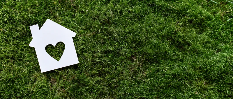White paper house cutout with heart-shaped window on green mossy ground