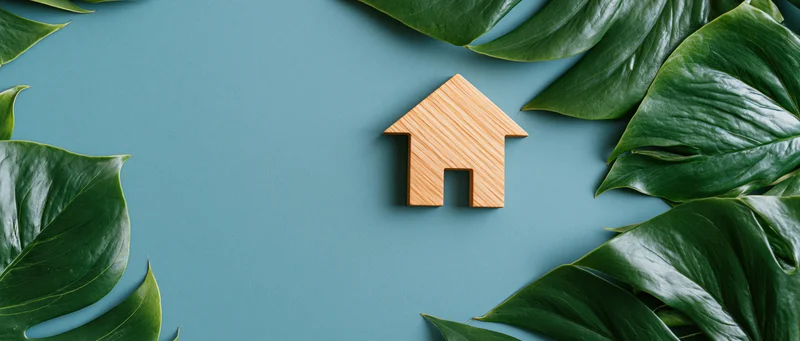 Wooden house cutout surrounded by monstera leaves on teal blue surface