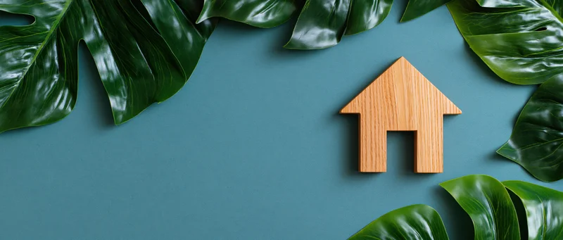 Wooden house cutout on teal surface surrounded by monstera leaves in flat lay arrangement