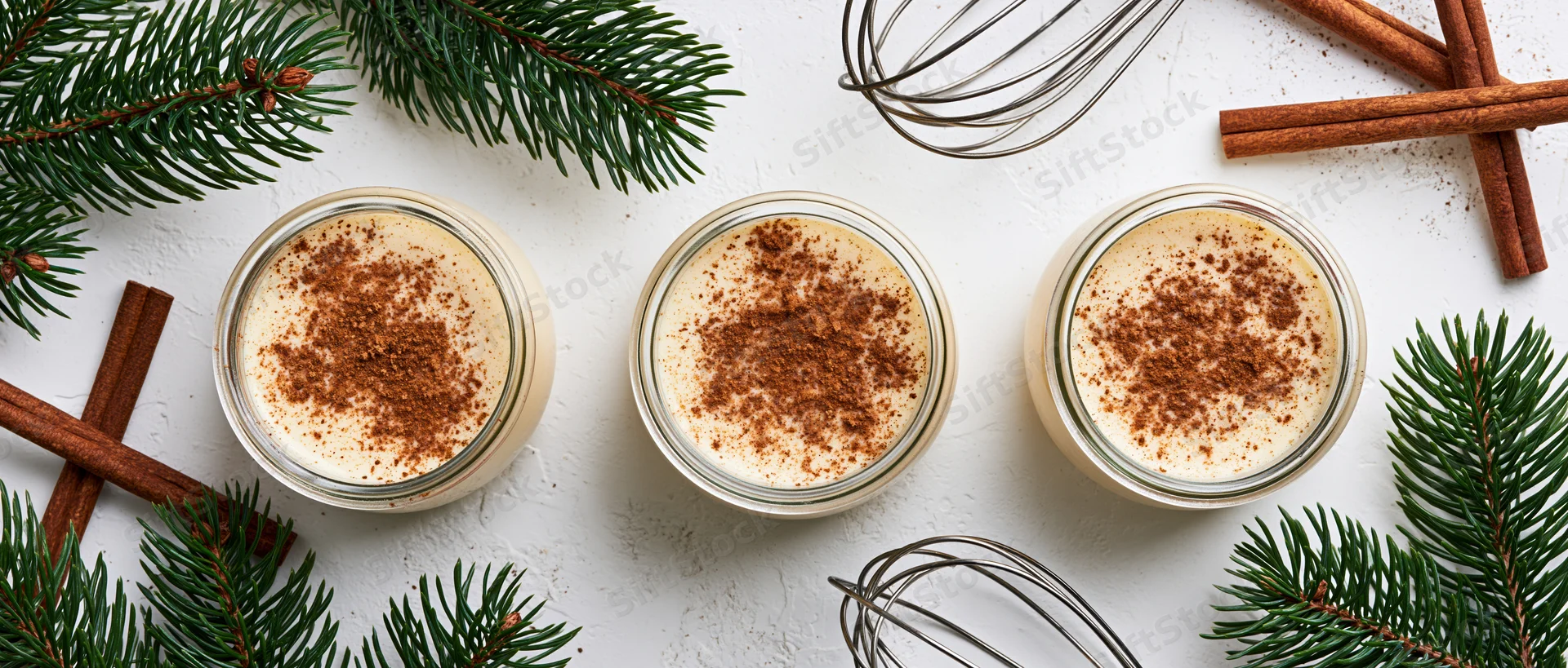Three glass jars of creamy eggnog dusted with cinnamon arranged on a white surface with pine branches and cinnamon sticks