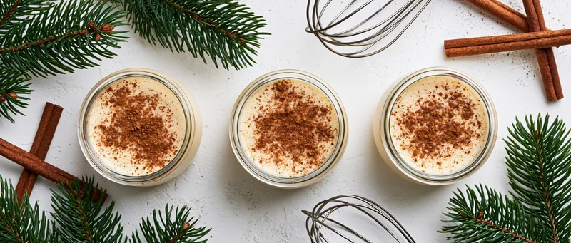 Three glass jars of creamy eggnog dusted with cinnamon arranged on a white surface with pine branches and cinnamon sticks