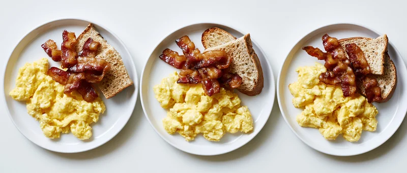 Three plates with scrambled eggs, bacon, and toast arranged side by side on a white surface