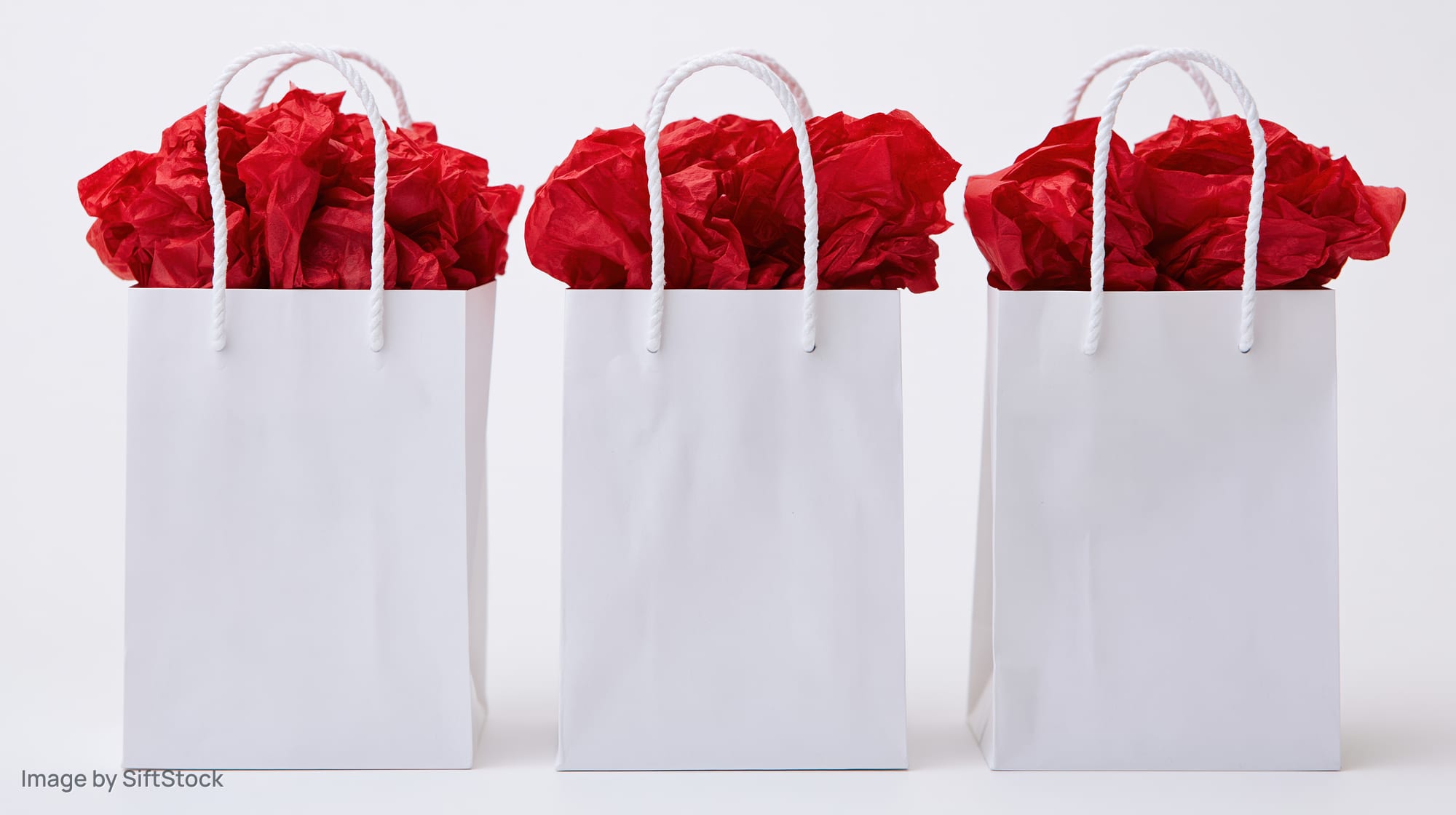 Three white paper gift bags filled with red tissue paper standing in a row on a white surface