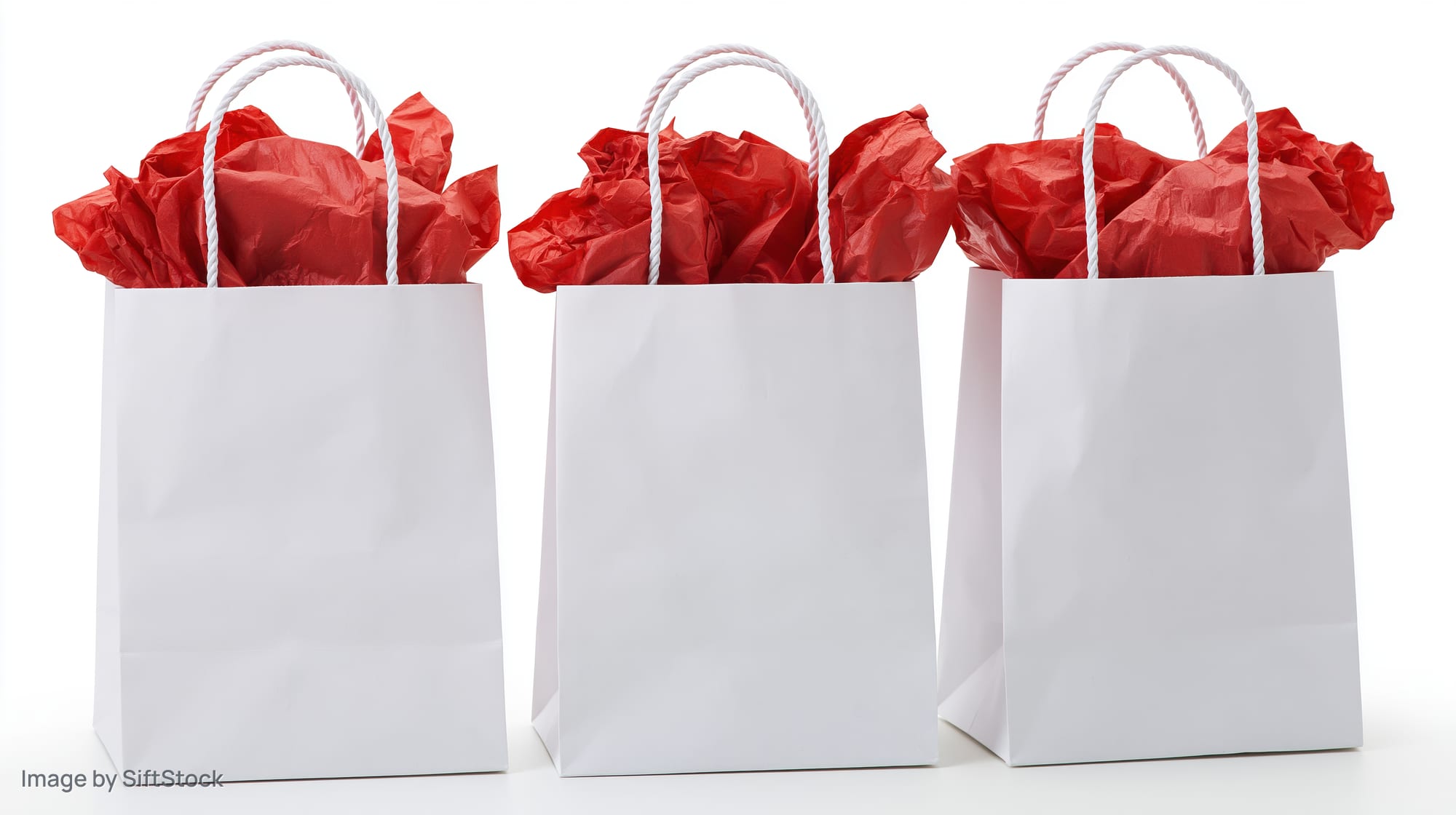 Three white paper gift bags filled with red tissue paper standing in a row on white background