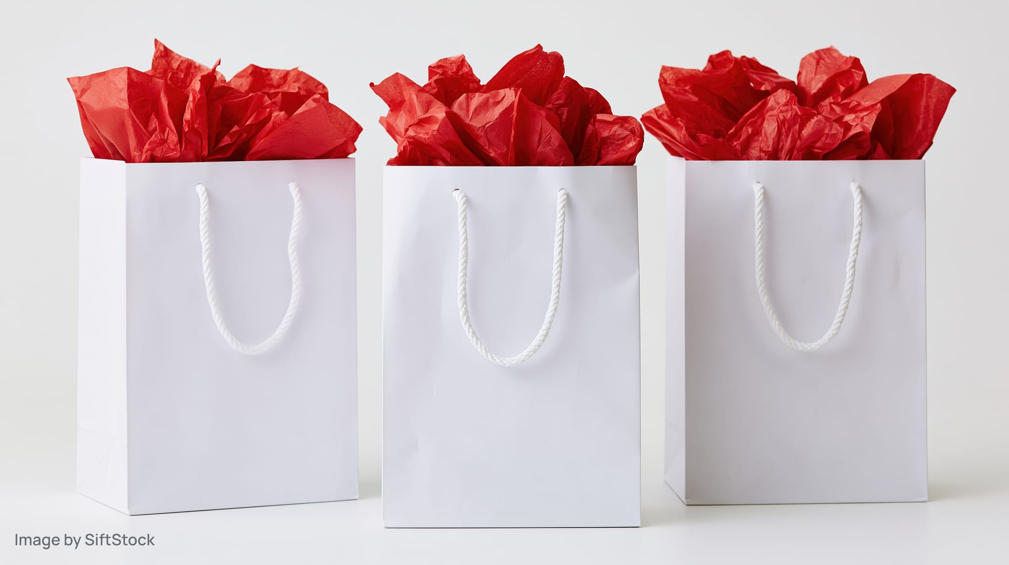 Three white paper gift bags with red tissue paper standing in a row on a white surface