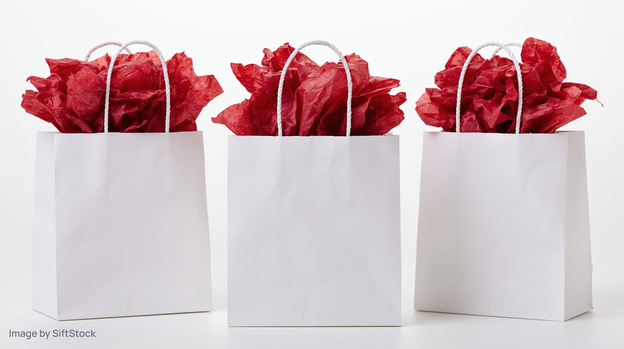 Three white paper gift bags filled with red tissue paper standing in a row on a white surface