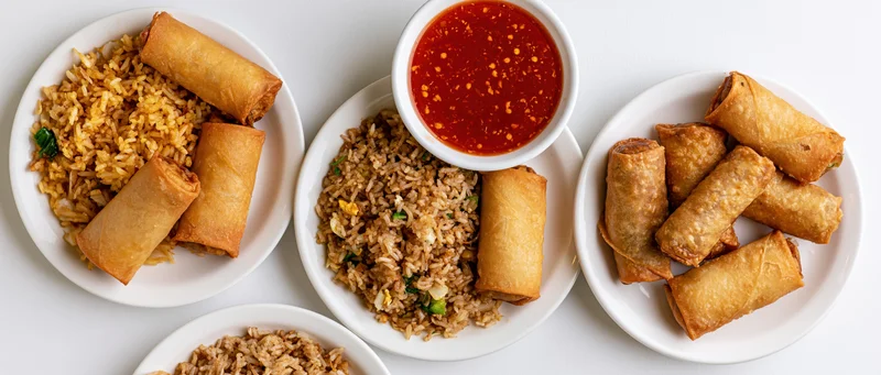 Three white plates with spring rolls and fried rice arranged side by side on a white surface