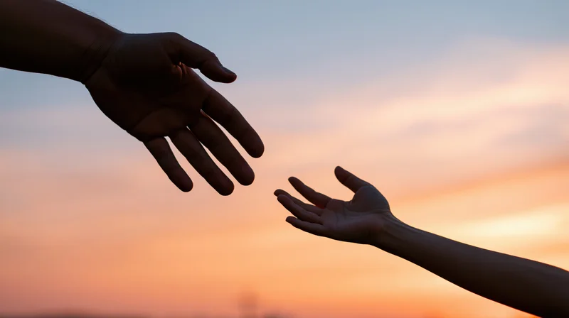 Two hands silhouetted reaching toward each other against a sunset sky
