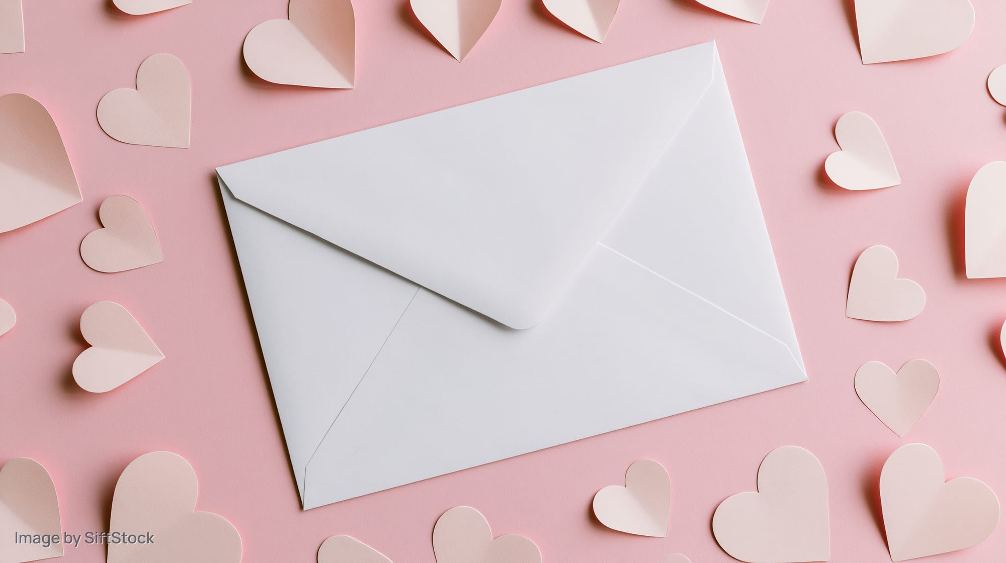 White envelope on pastel pink surface surrounded by paper hearts in flat lay arrangement