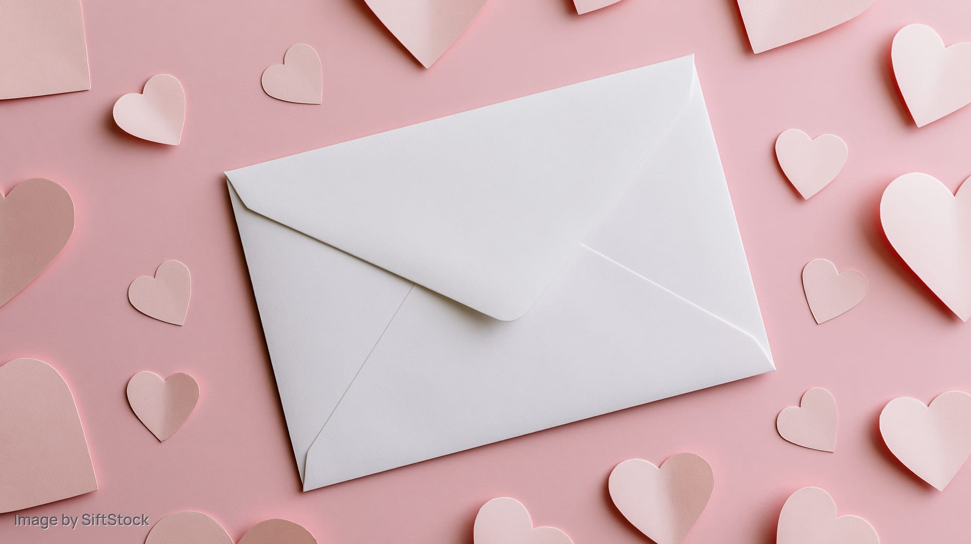 White envelope on pastel pink surface surrounded by paper hearts in flat lay arrangement