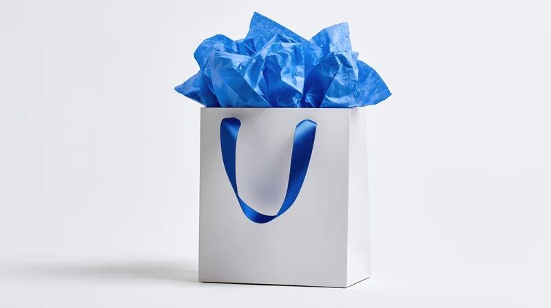 White matte gift bag with blue satin ribbon handles filled with crumpled blue tissue paper on plain white background