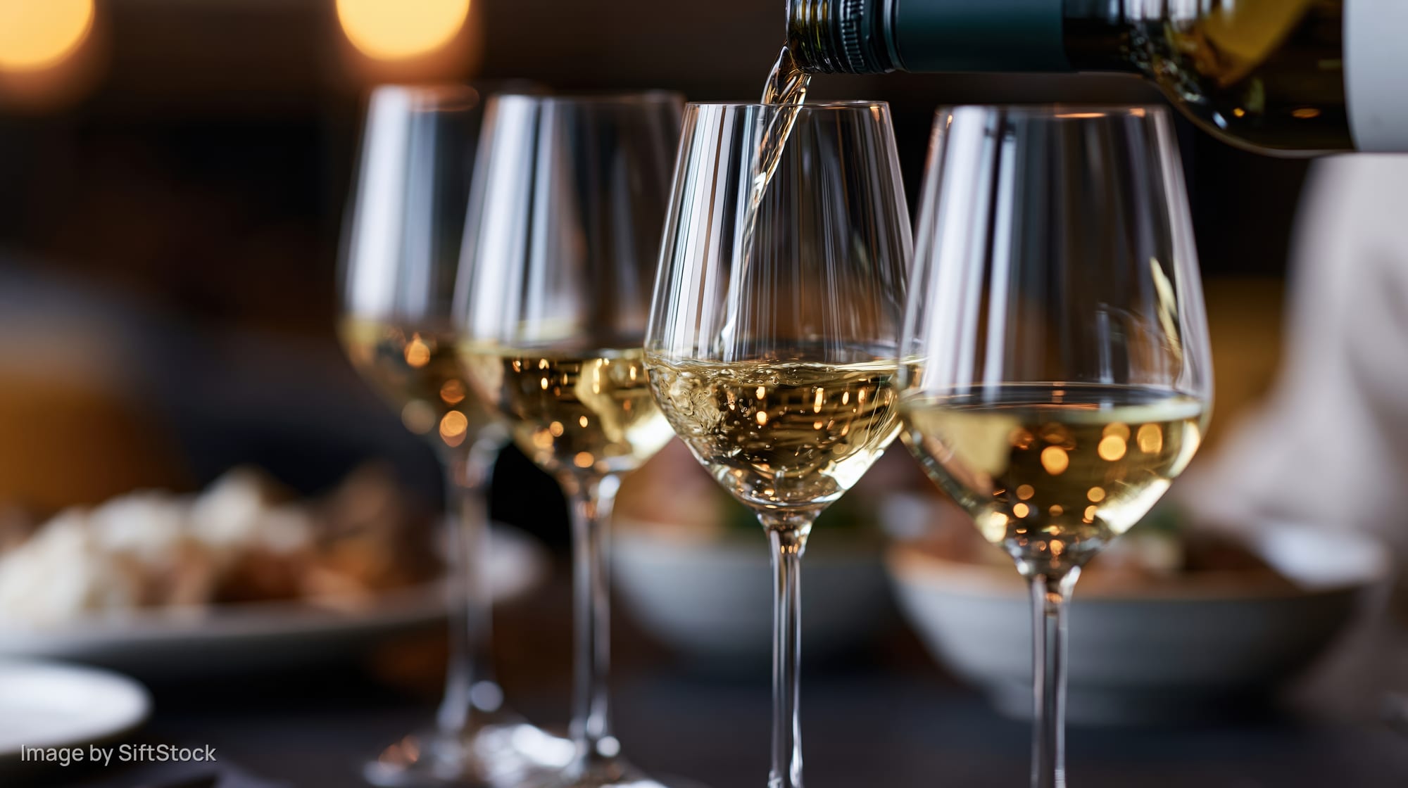 White wine being poured from a bottle into one of four glasses on a dining table