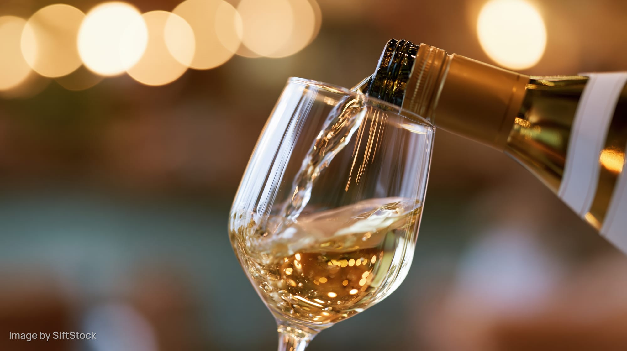 White wine being poured from a gold-labeled bottle into a clear glass at a dimly lit indoor setting
