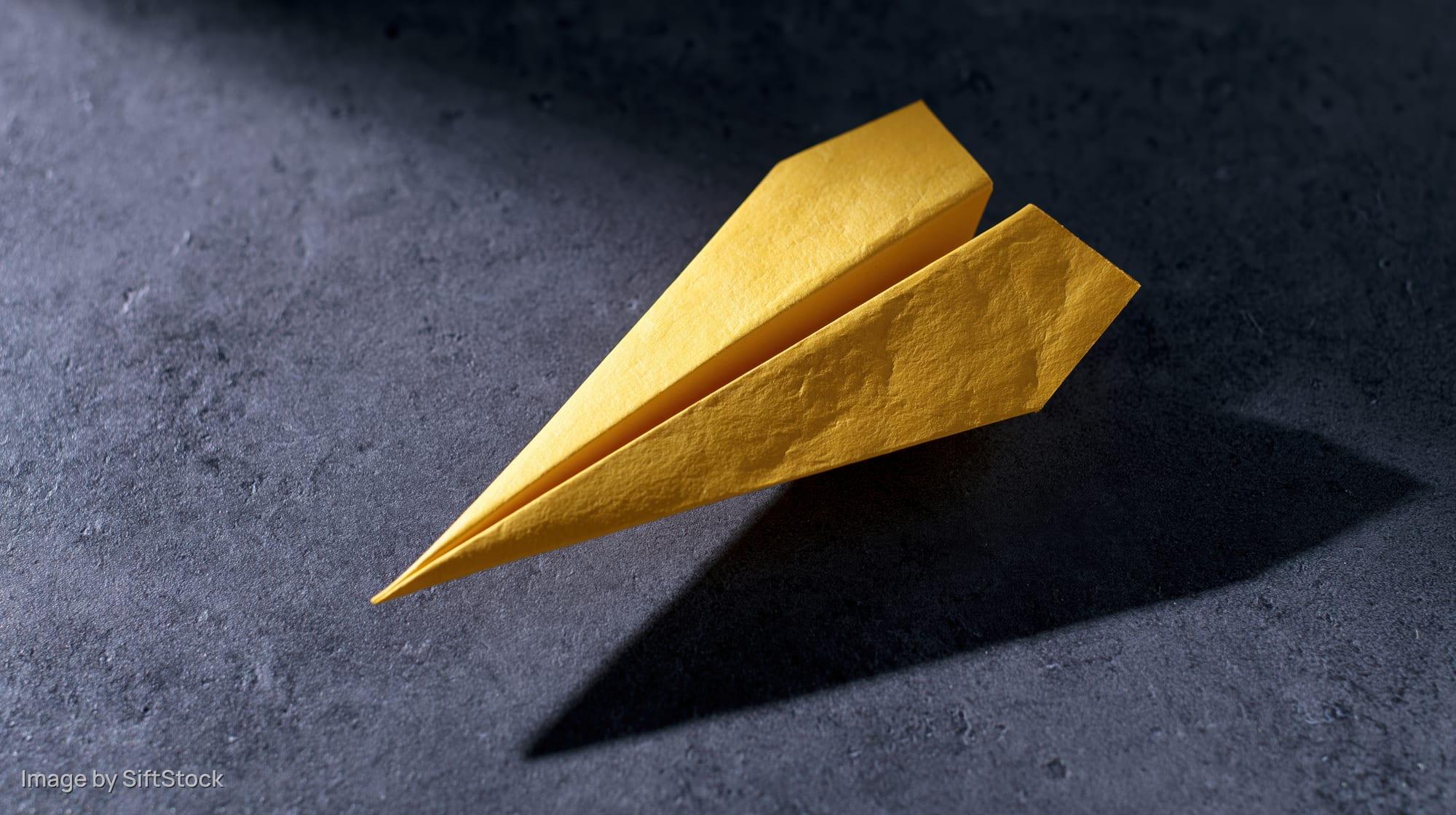 Yellow paper airplane folded and resting on dark textured surface casting sharp shadow