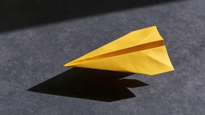 Yellow paper airplane resting on dark surface casting sharp shadow