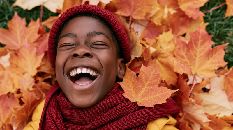 Young black child laughing with eyes closed while lying in a pile of autumn maple leaves