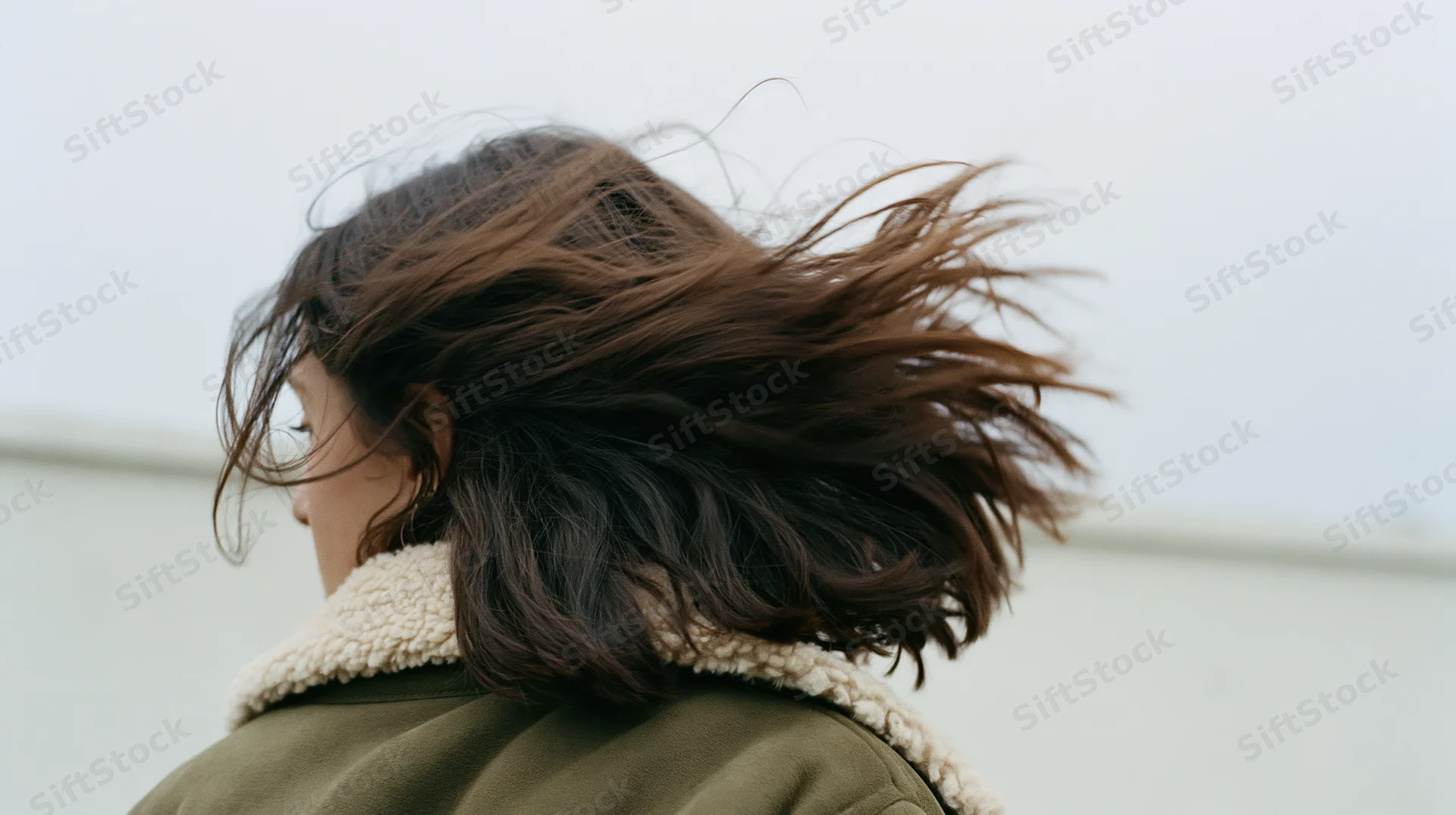 Woman from behind with dark brown hair blowing in wind wearing olive coat with shearling collar