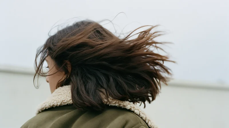 Woman from behind with dark brown hair blowing in wind wearing olive coat with shearling collar