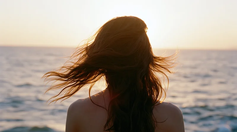 Woman seen from behind with long hair blowing in the wind at ocean's edge during sunset